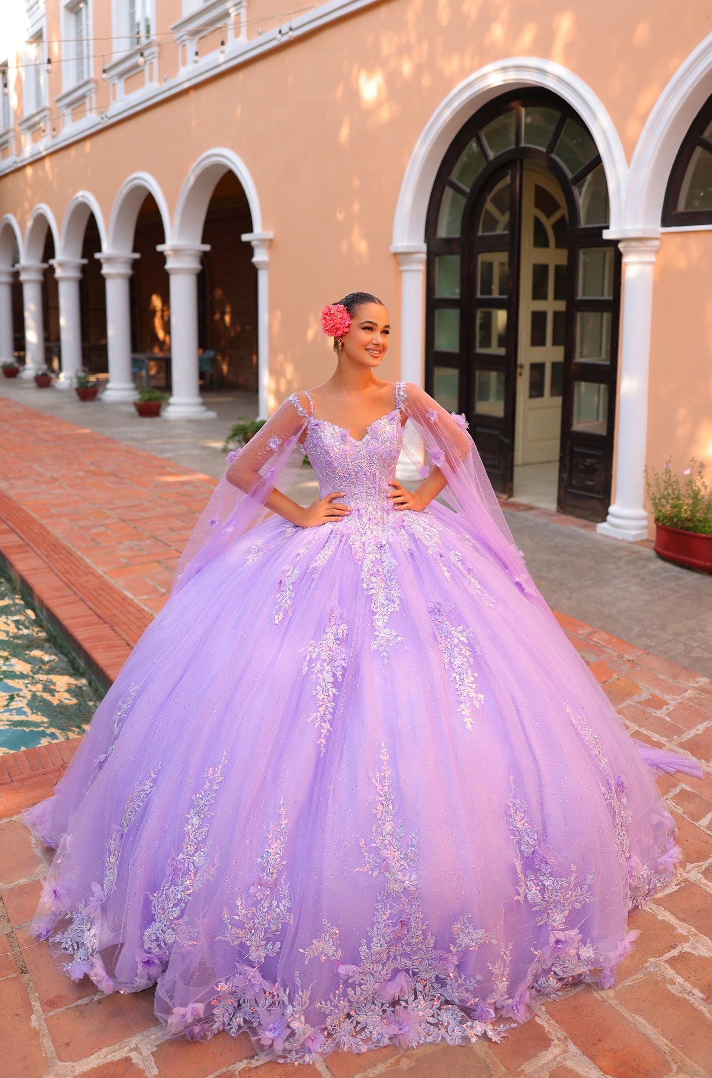 A woman wears a voluminous lilac quinceañera gown with lace details, posing in an arched colonnade.