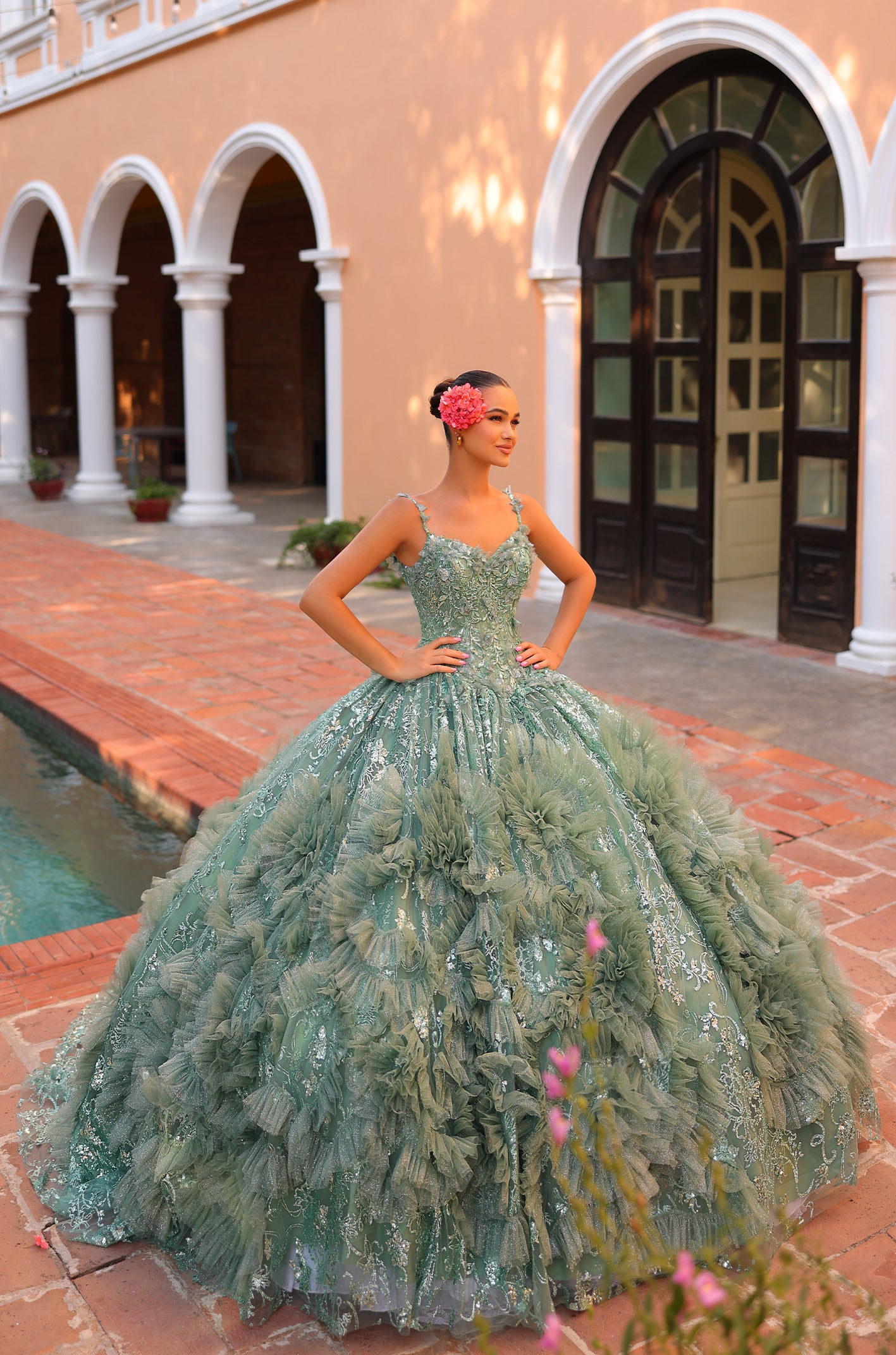 A sage green quinceañera dress with ruffles and sparkles, worn by a woman posing in an arched courtyard.