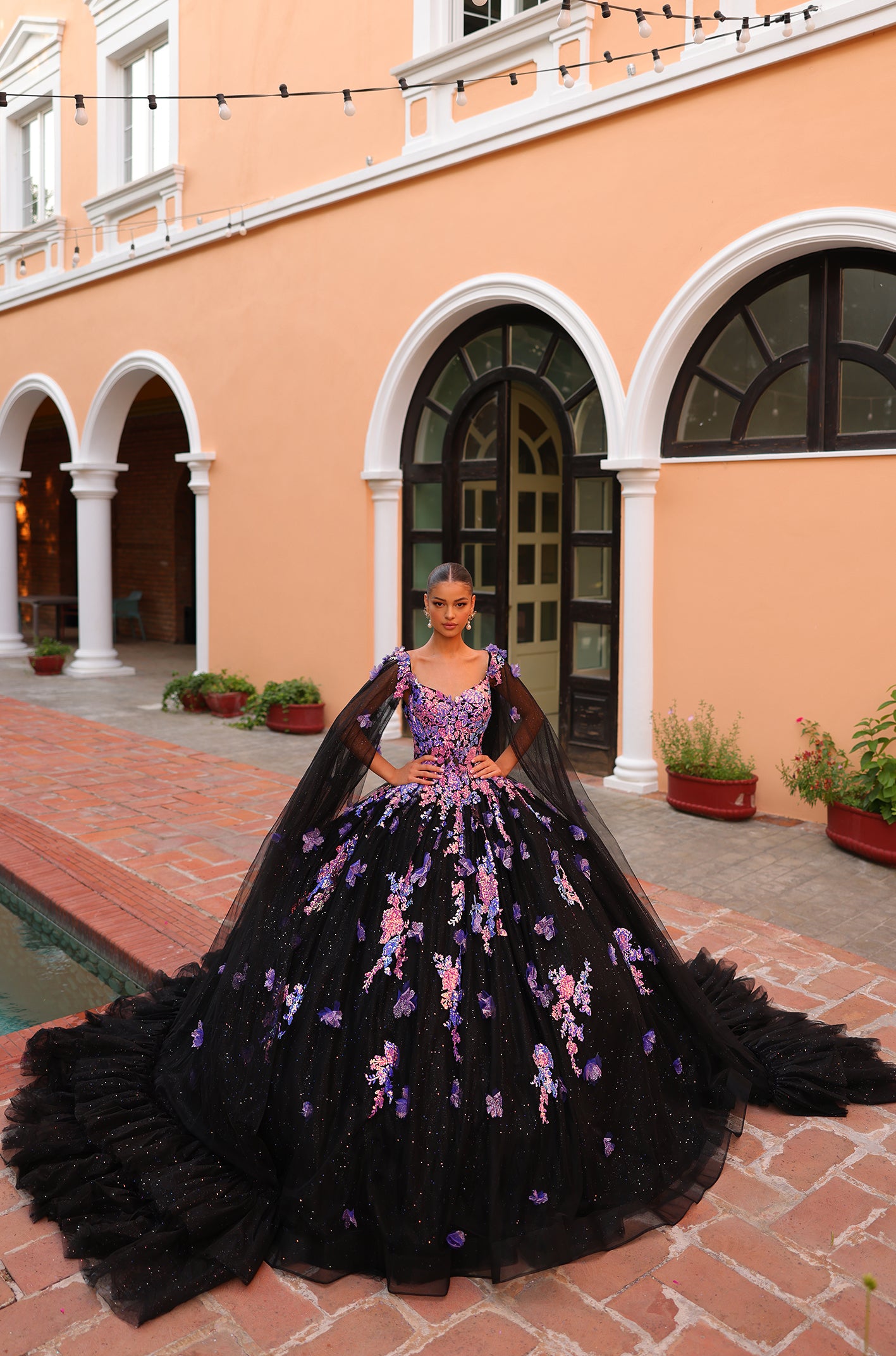 A Black/Multi ball gown with purple floral embroidery stands on a brick courtyard with arched windows and string lights.