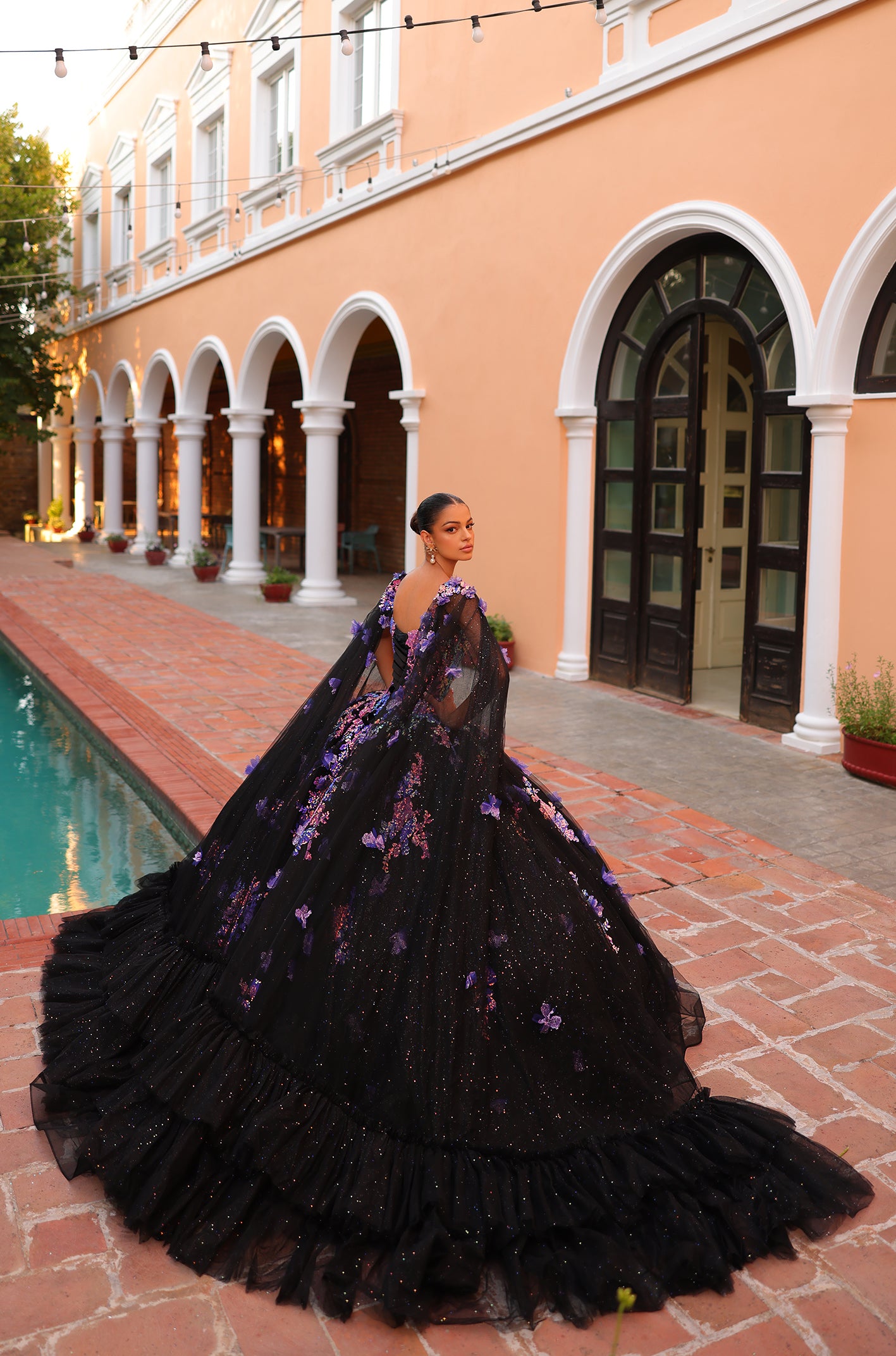 A person in a black sparkling ball gown with purple floral accents stands by a pool near a peach-colored building.