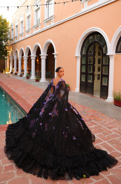 A person in a black sparkling ball gown with purple floral accents stands by a pool near a peach-colored building.