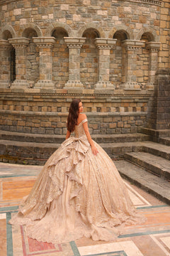 A woman in an elaborate, off-shoulder champagne lace ball gown poses before a stone architectural backdrop.