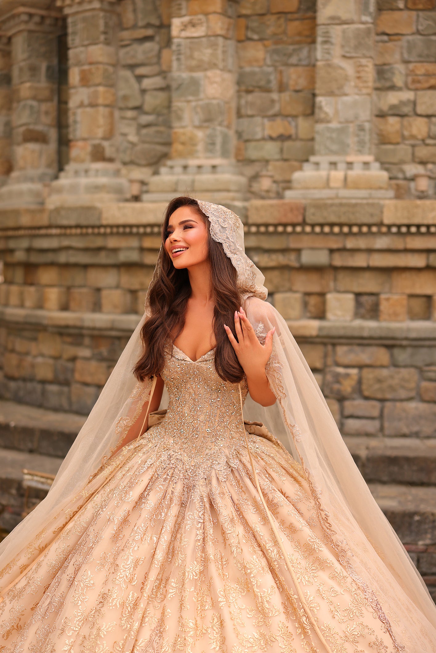 A bride in a sparkling gold ballgown and lace veil smiles joyfully in front of an ornate stone architectural backdrop.