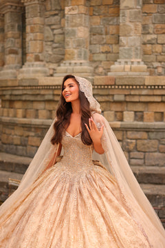 A bride in a sparkling gold ballgown and lace veil smiles joyfully in front of an ornate stone architectural backdrop.