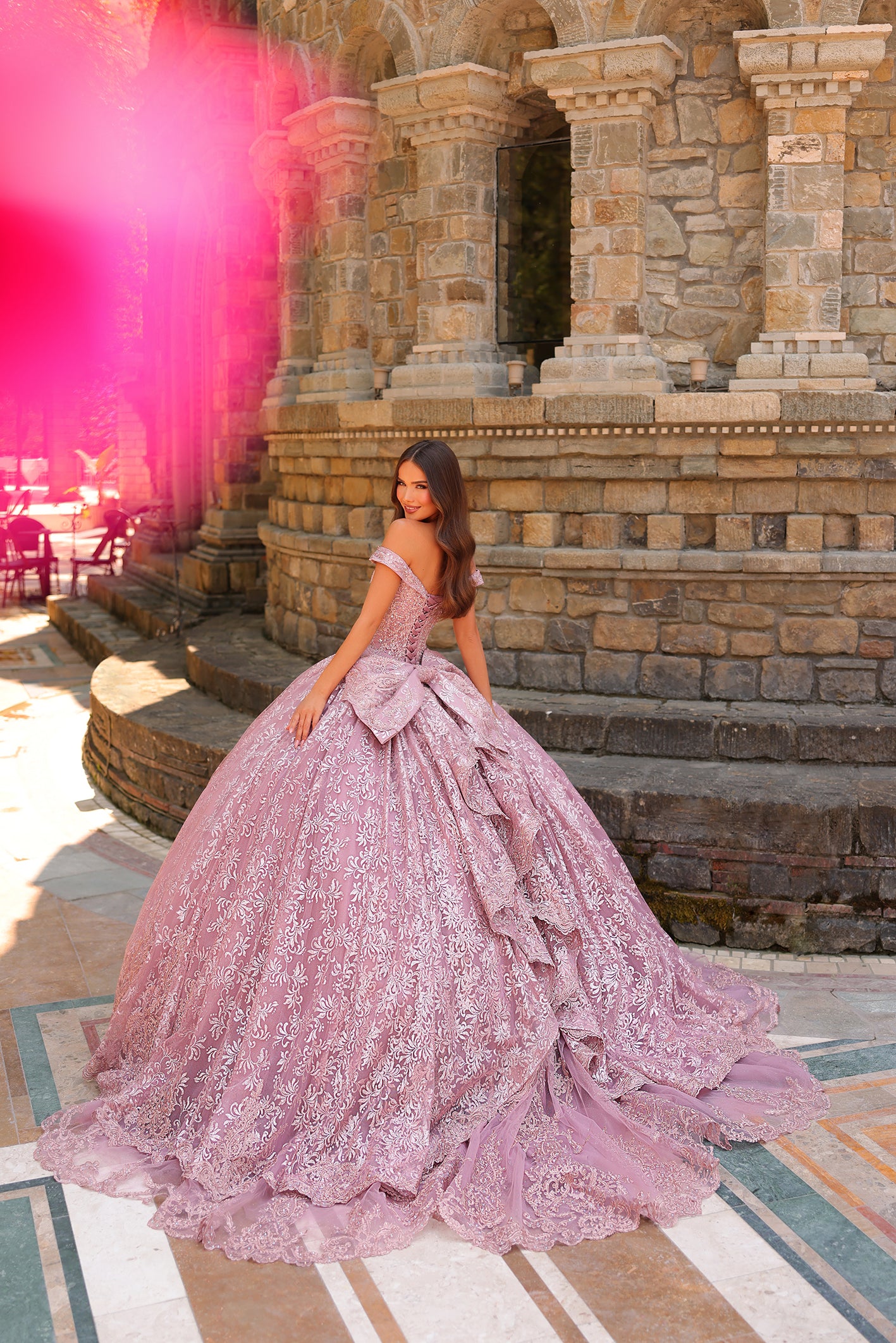A young woman wears a lavender lace ball gown with an off-shoulder design, posing on stone steps.