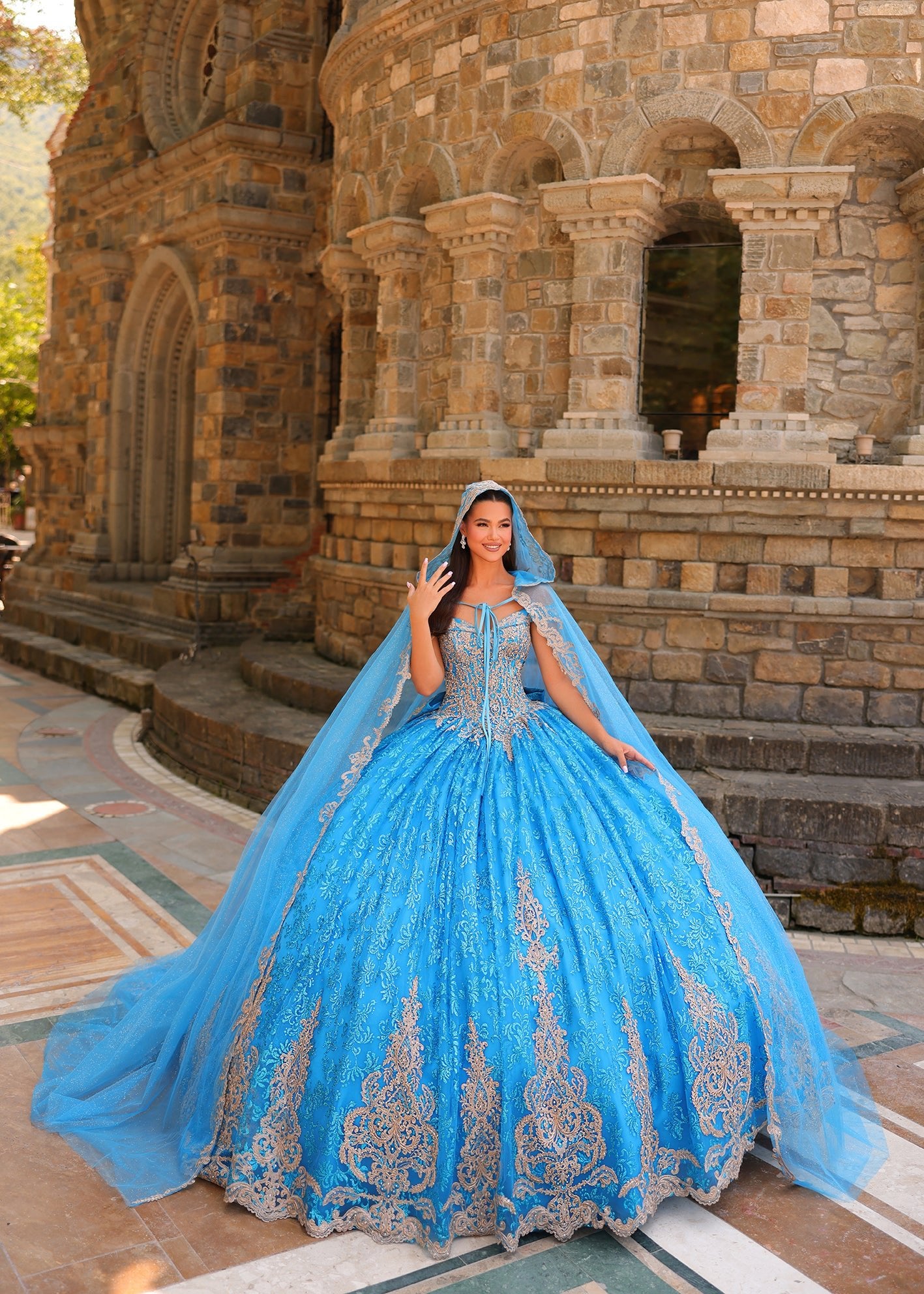 A woman in a voluminous blue ball gown with silver embroidery poses before a stone medieval-style building.