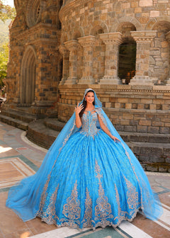 A woman in a voluminous blue ball gown with silver embroidery poses before a stone medieval-style building.