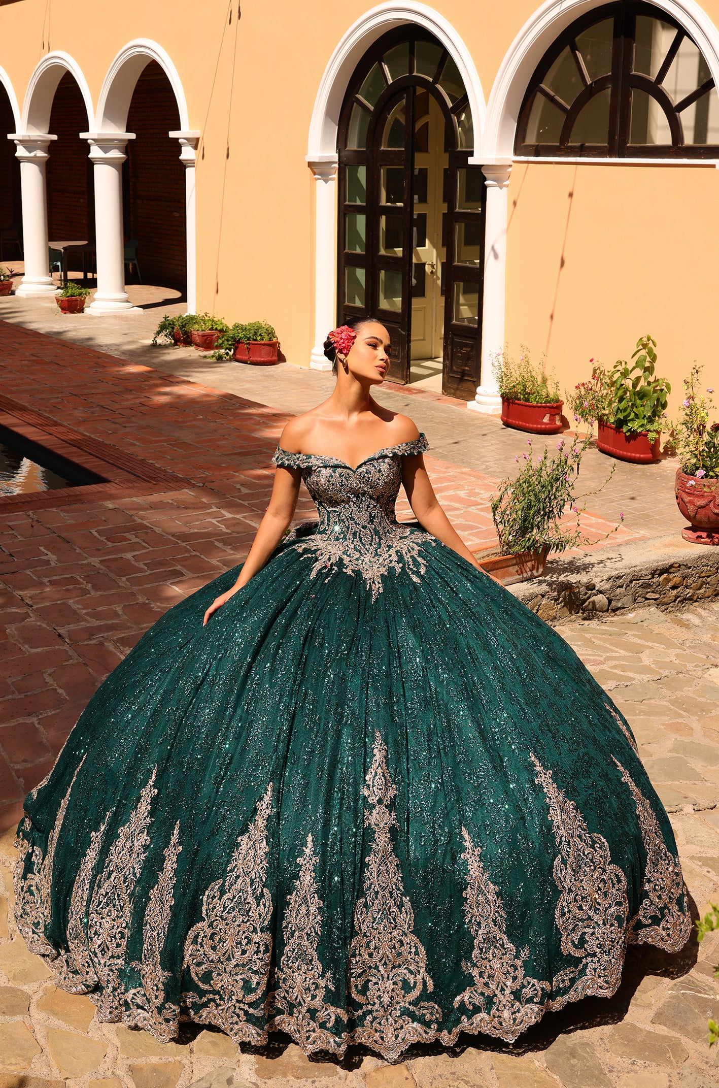 A woman wears a sparkling emerald/gold off-shoulder quinceañera ball gown with intricate lace details in an arched.