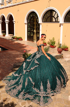 A woman wears an emerald green, off-shoulder ball gown with silver lace, posing in a sunlit courtyard with arched windows.