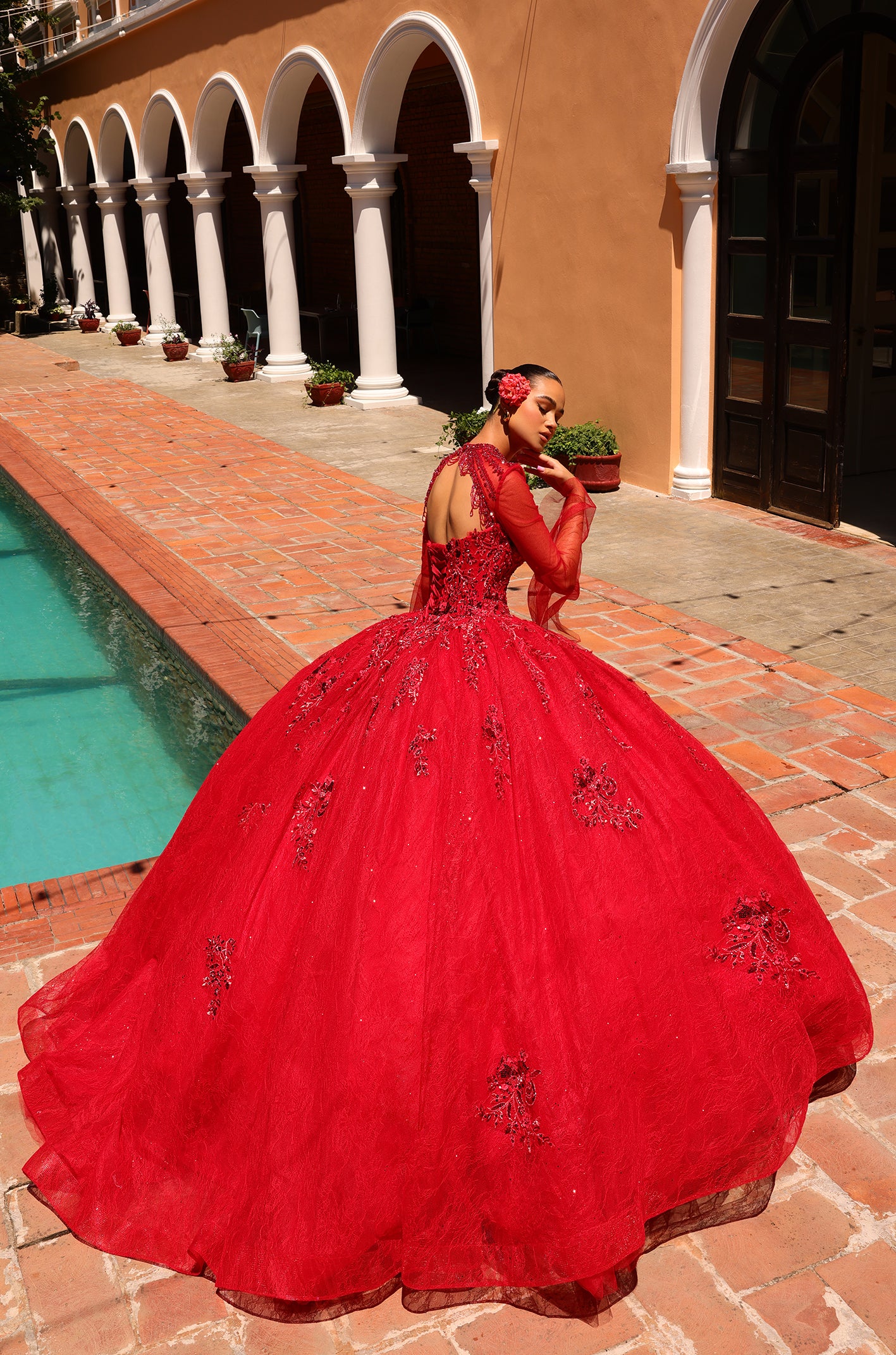 A woman in a vibrant red ballgown poses elegantly by a pool near white-columned arches.