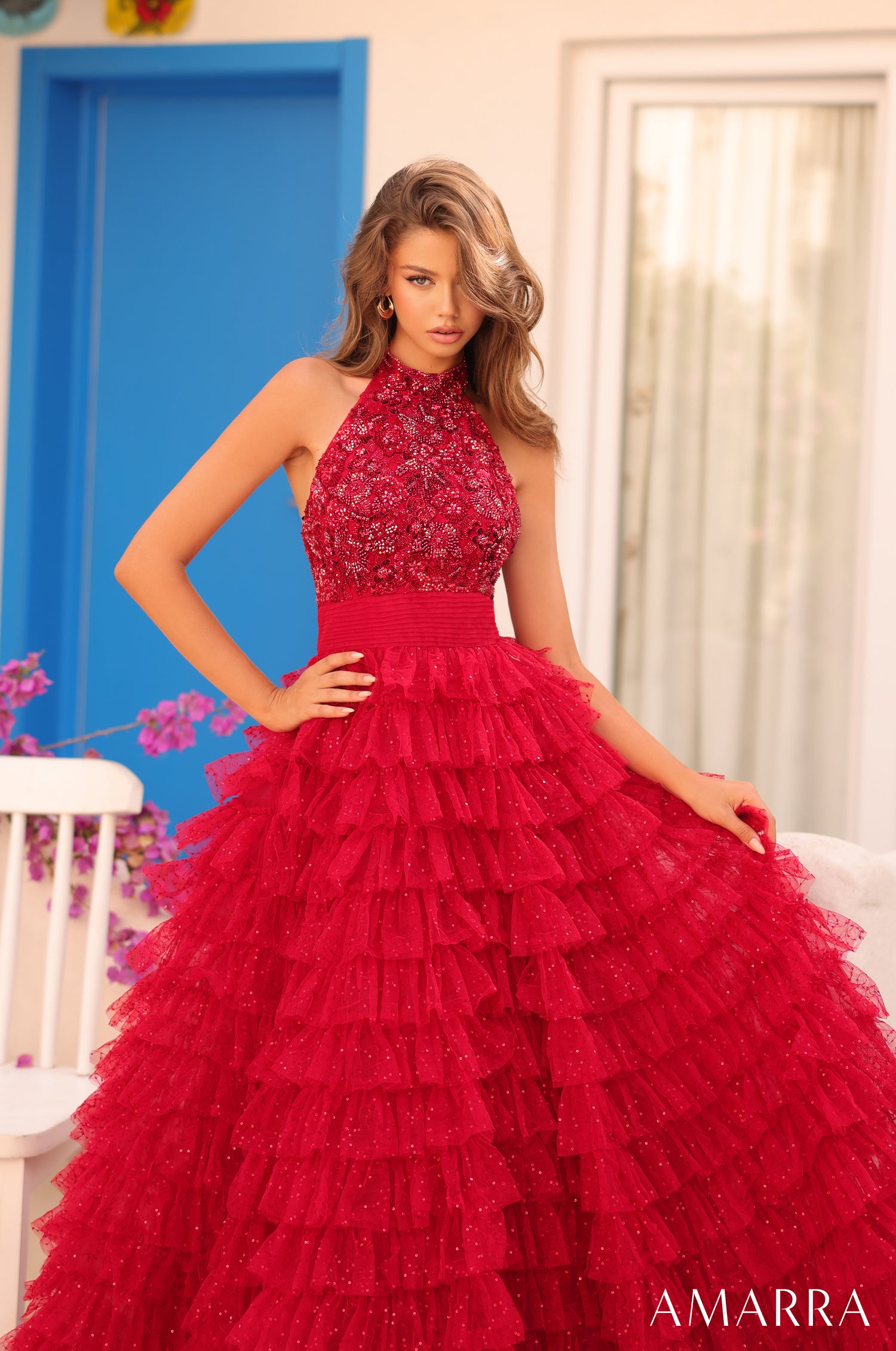 Model wears a red tiered, sequined halter ball gown with a fitted bodice against a blue and white backdrop.