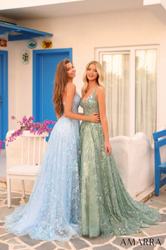 Two women wear elegant floor-length embroidered gowns in blue and green, posing together in a Mediterranean-style setting.