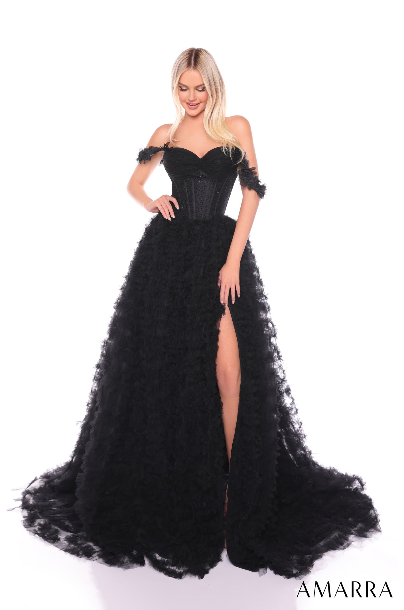 A blonde woman wears a black ruffled off-shoulder ball gown with a high slit on a white background.