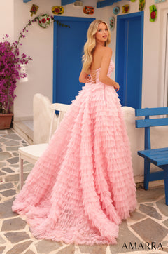 A blonde model wears a pink ruffled ballgown with a corset top, standing in a blue-and-white Mediterranean-style setting.