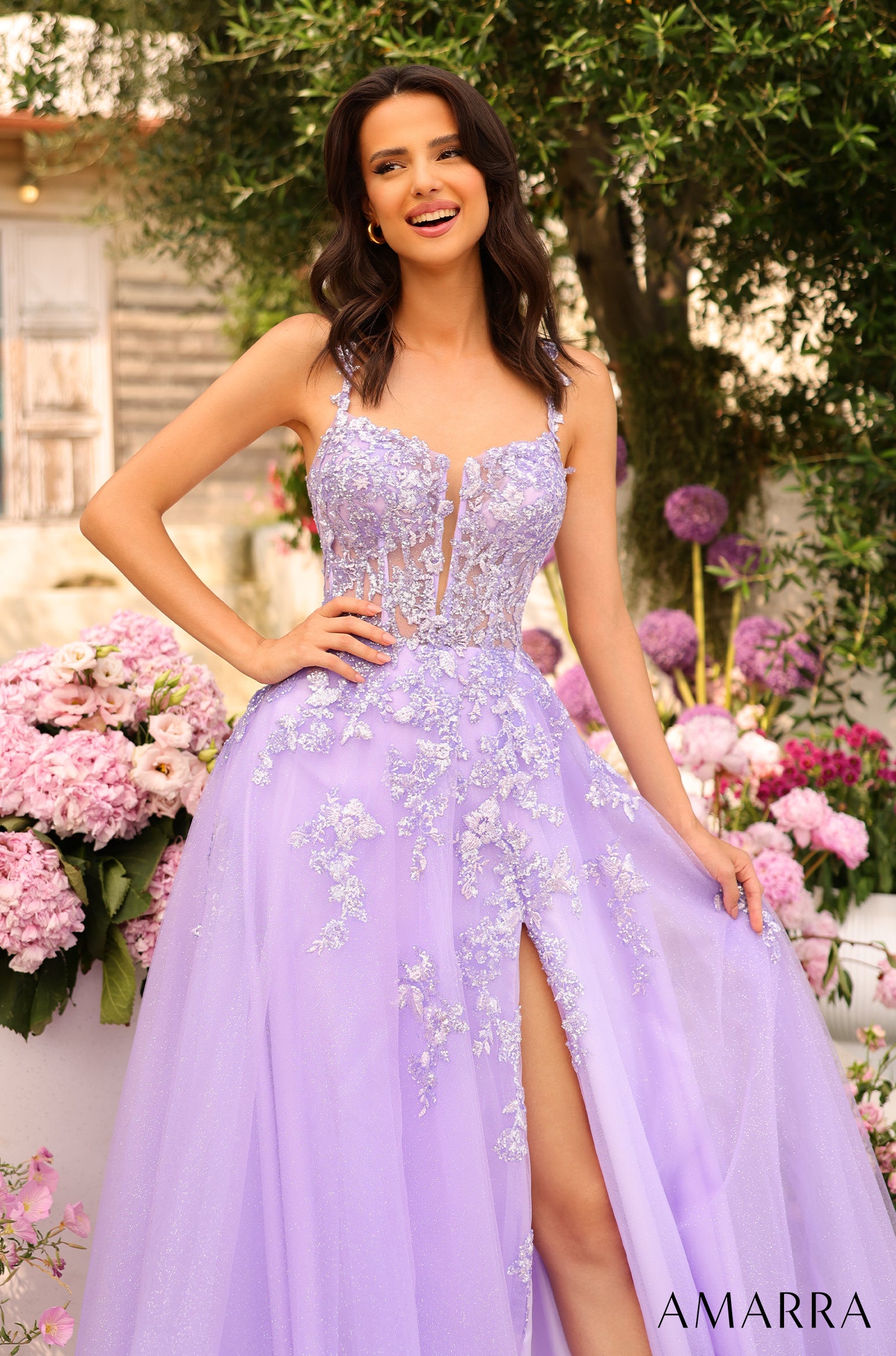A woman in a lavender ball gown with floral embellishments smiles in a garden of pink and purple flowers.