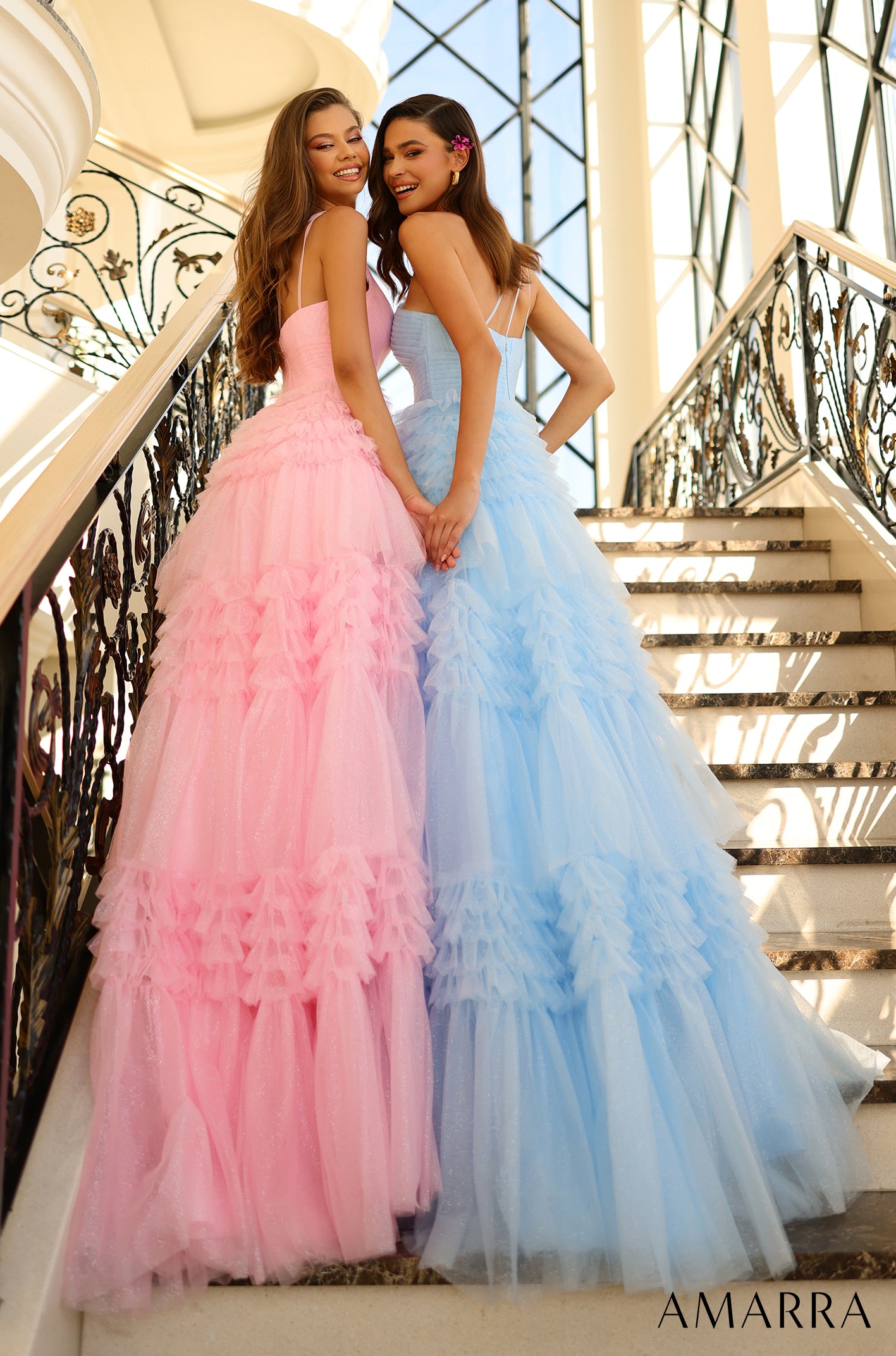 Two women in ruffled tulle gowns, one pink and one blue, stand together on an ornate staircase.