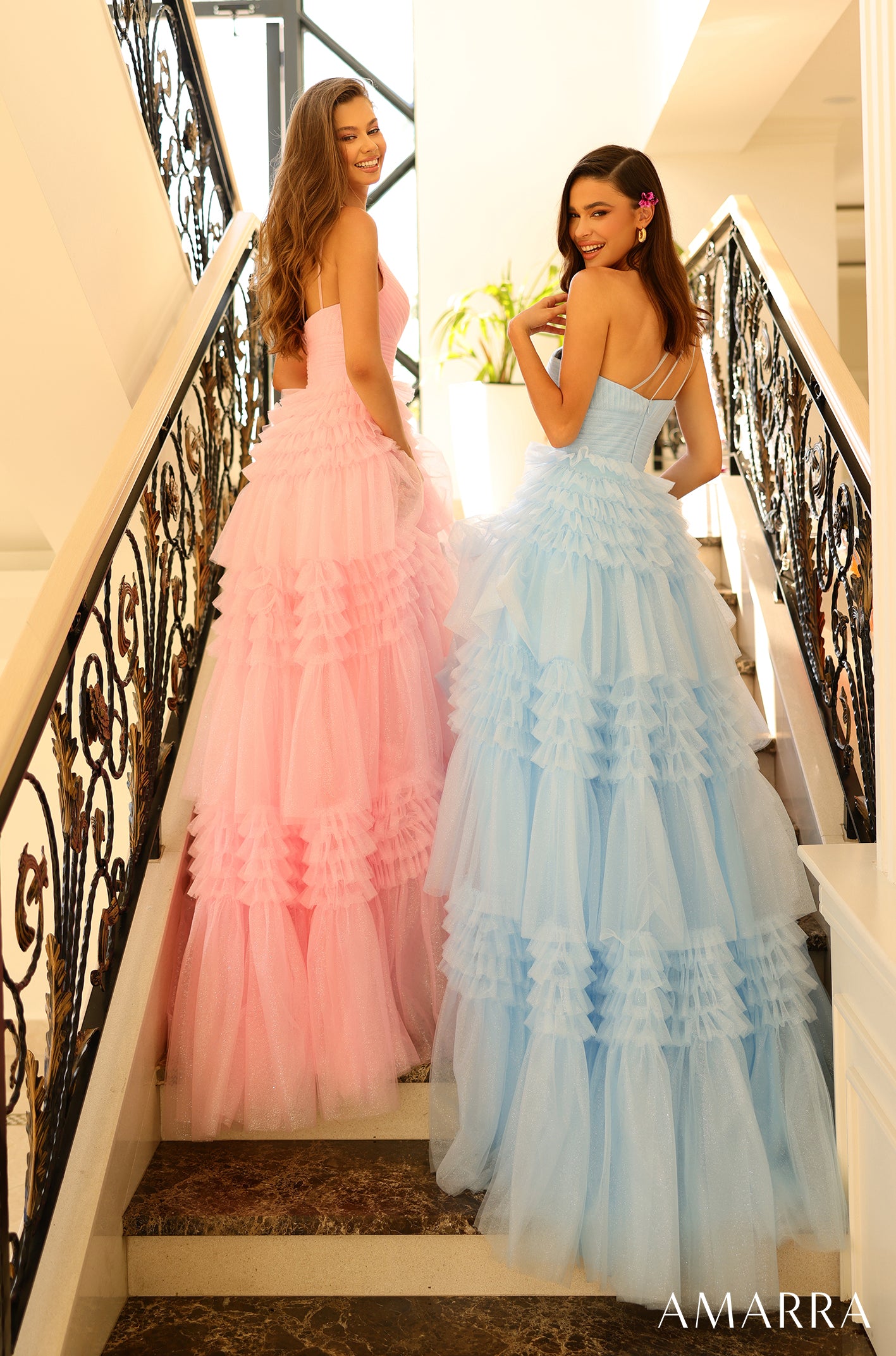 Two women in ruffled tulle gowns - pink and blue - pose on an ornate staircase with decorative wrought-iron railings.