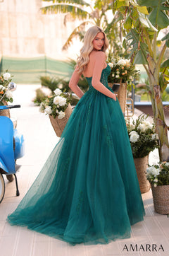 A model wears a teal tulle ball gown with floral embellishments, standing among flowers and palm trees.