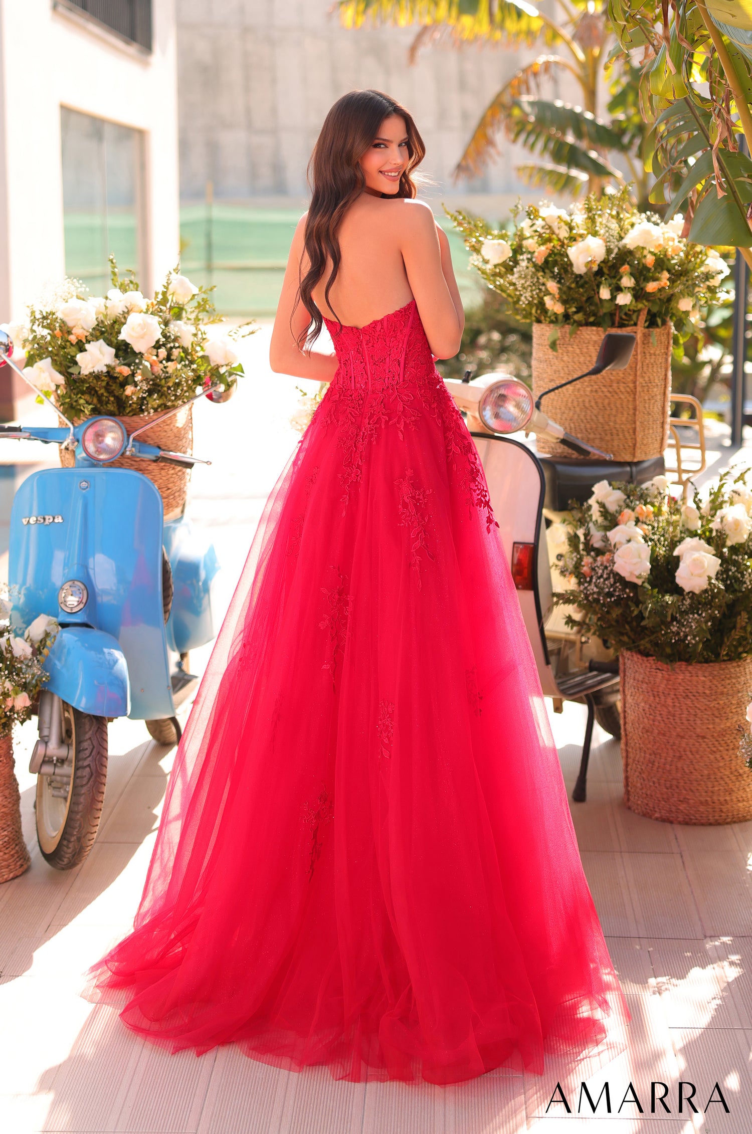 A woman in a vibrant pink ball gown poses near a blue Vespa with flower arrangements.