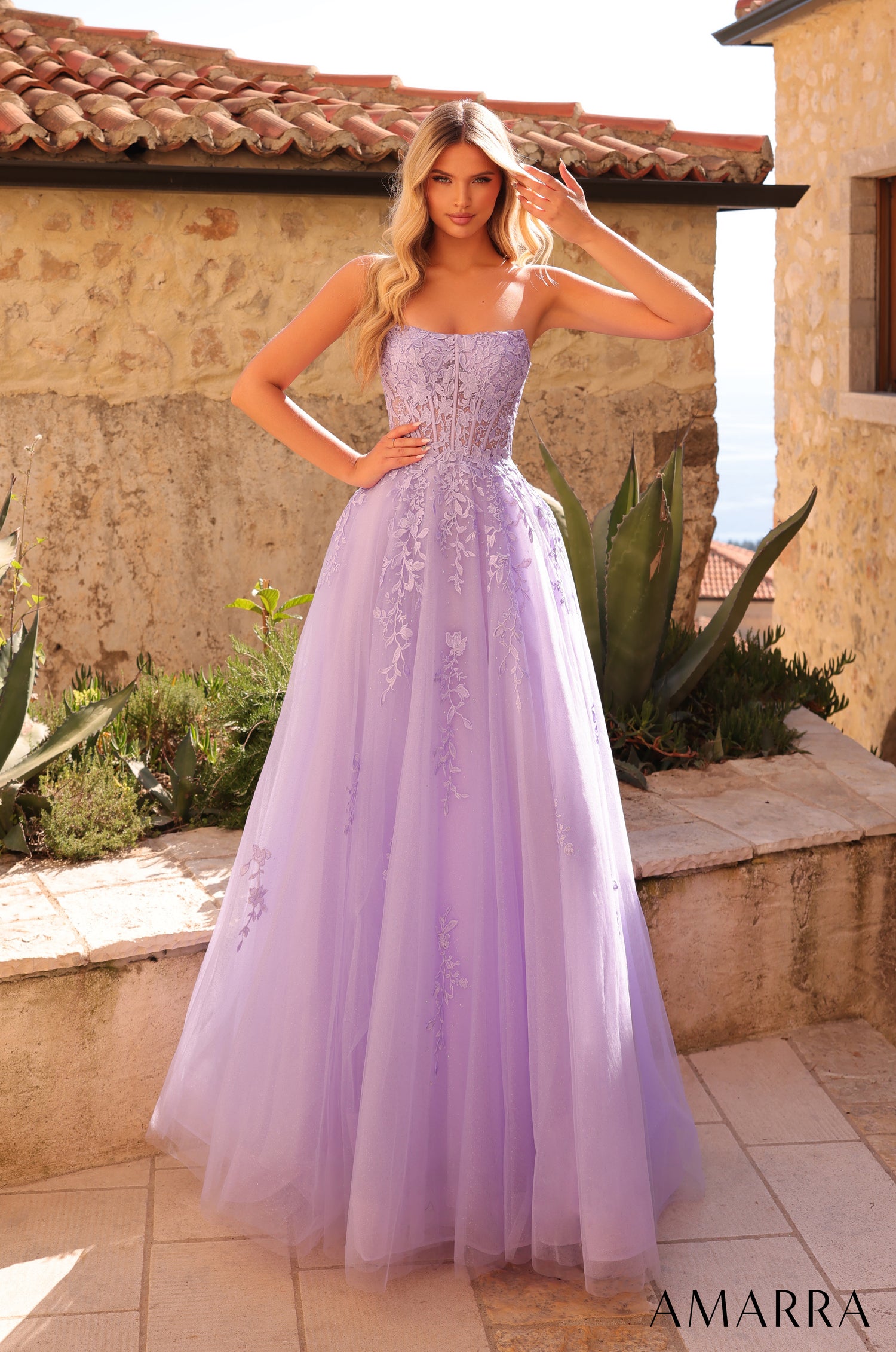 A model wears a strapless, floor-length lilac tulle ball gown with floral embroidery in a Mediterranean-style setting.