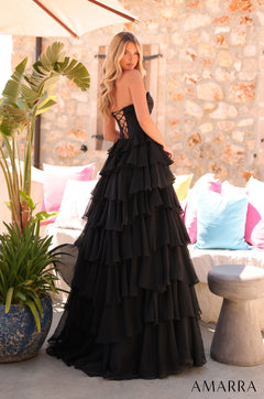 A model wears a black tiered ballgown with a corset top, standing in a sunlit room with colorful pillows.