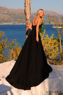 A blonde model wears a black strapless ball gown against a scenic ocean and mountain backdrop.