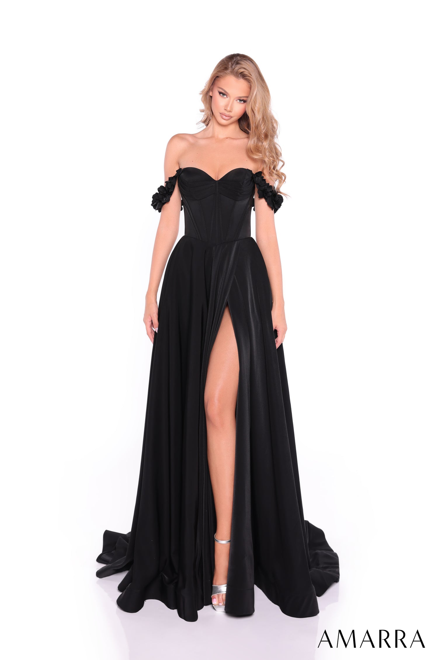 A woman wears a black off-shoulder gown with a high slit and ruffled sleeves, standing on a white background.