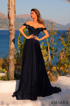 A woman in a navy off-shoulder gown stands against a scenic ocean and mountain backdrop with foliage.