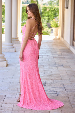 A woman wears a long, bright pink sequined gown with a criss-cross back, standing on stone tiles near white columns.