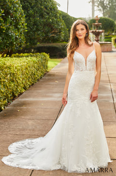 A bride in a white lace mermaid wedding dress stands on a garden path with a fountain in the background.