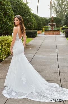 A bride in a white lace mermaid wedding dress poses by a fountain with manicured gardens behind her.
