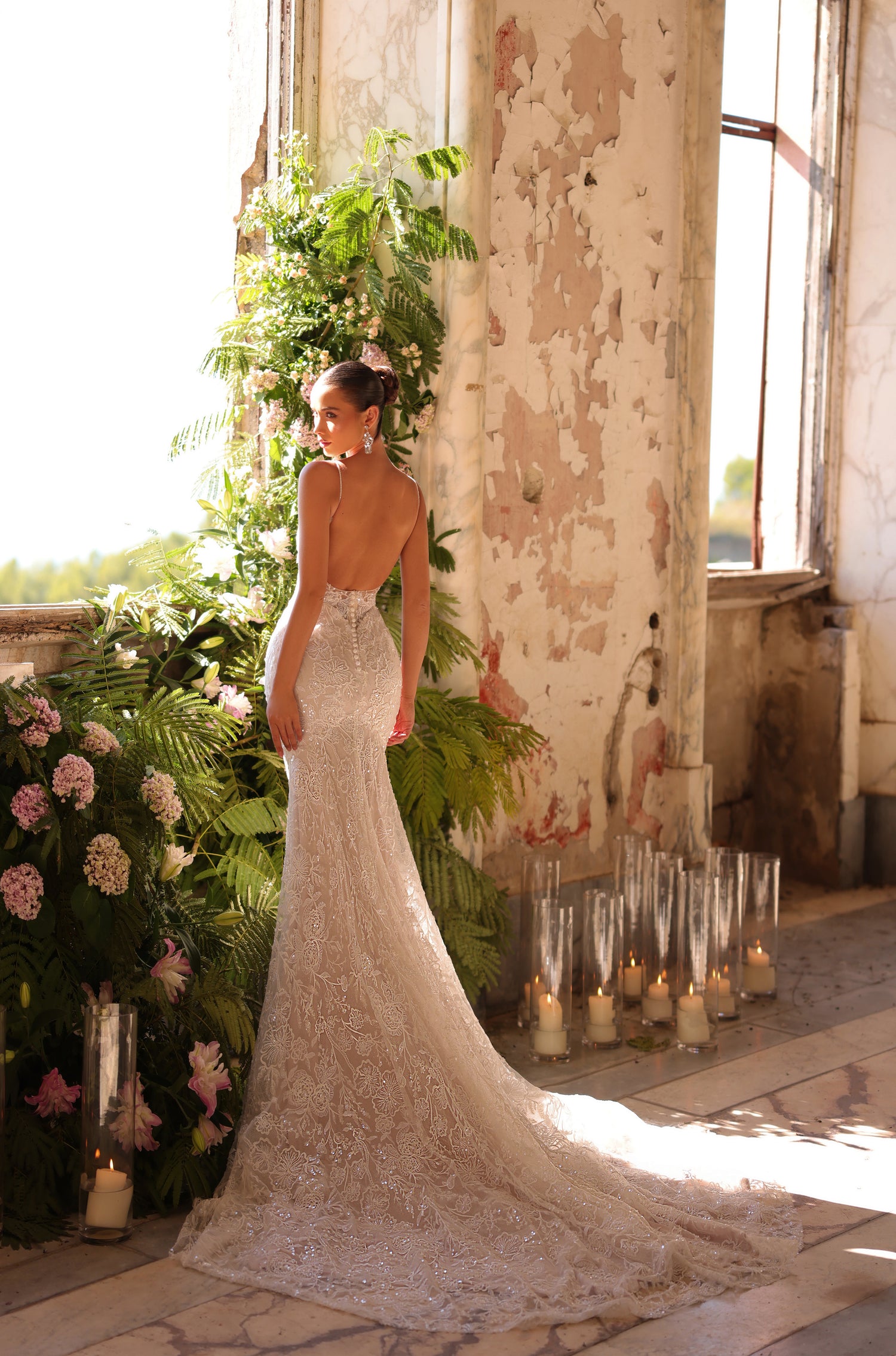 A bride in a backless lace mermaid wedding gown stands by candles and lush greenery in a weathered room.