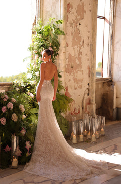 A bride in a backless lace mermaid wedding gown stands by candles and lush greenery in a weathered room.
