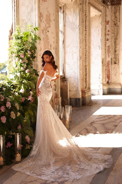 A bride in a lace mermaid wedding gown poses in a sunlit, vintage room with floral and candle decorations.
