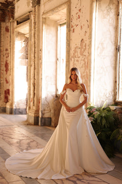 A bride in an off-shoulder white wedding gown poses in a sunlit, distressed interior with plants nearby.