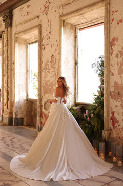 Bride in flowing white gown stands by window in distressed room with candles and greenery.