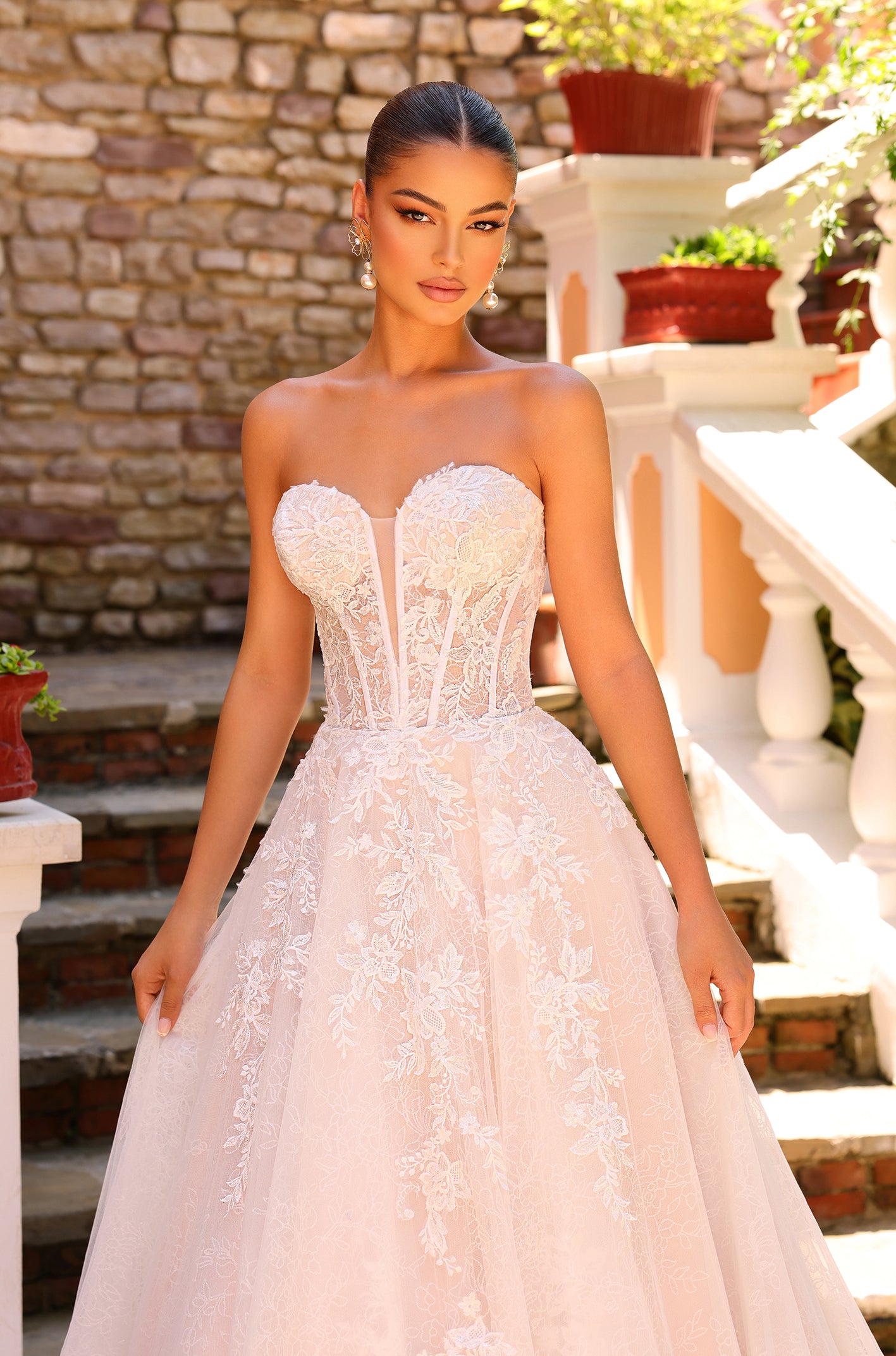 A bride stands in an elegant, strapless white lace ball gown against a stone wall and garden backdrop.