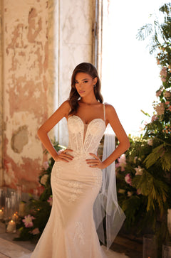 A bride in a fitted lace wedding gown stands confidently by flowers and candles in a vintage room.