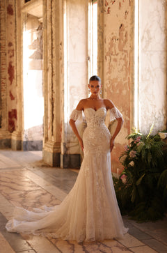 A bride in an off-shoulder lace mermaid wedding gown stands in a rustic, sunlit interior with plants nearby.