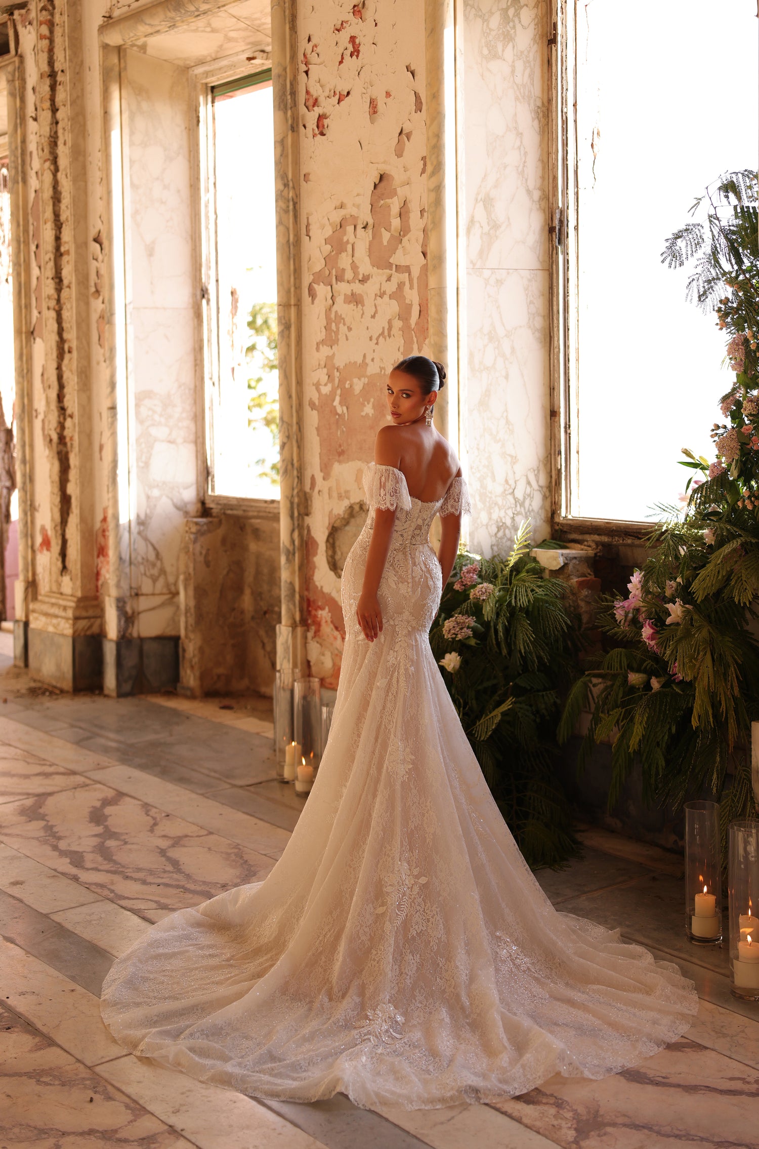 Bride in lace mermaid wedding gown poses by window with candles and tropical flowers.