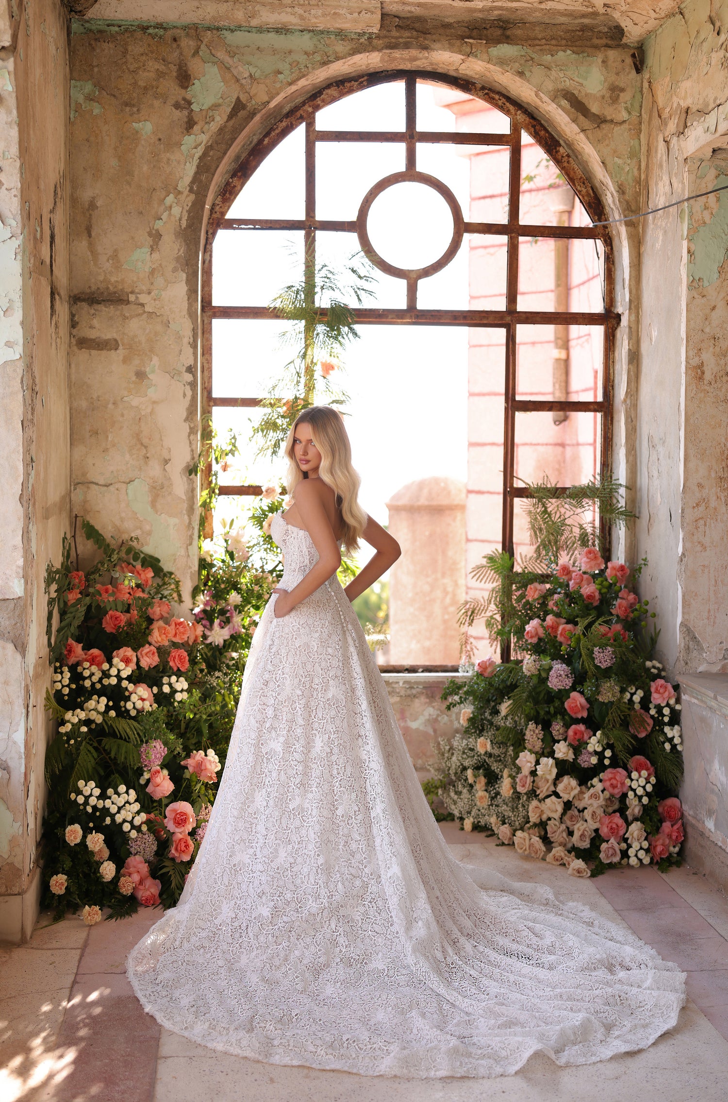 A bride in a lace wedding gown stands by an arched window, surrounded by pink and white floral arrangements.