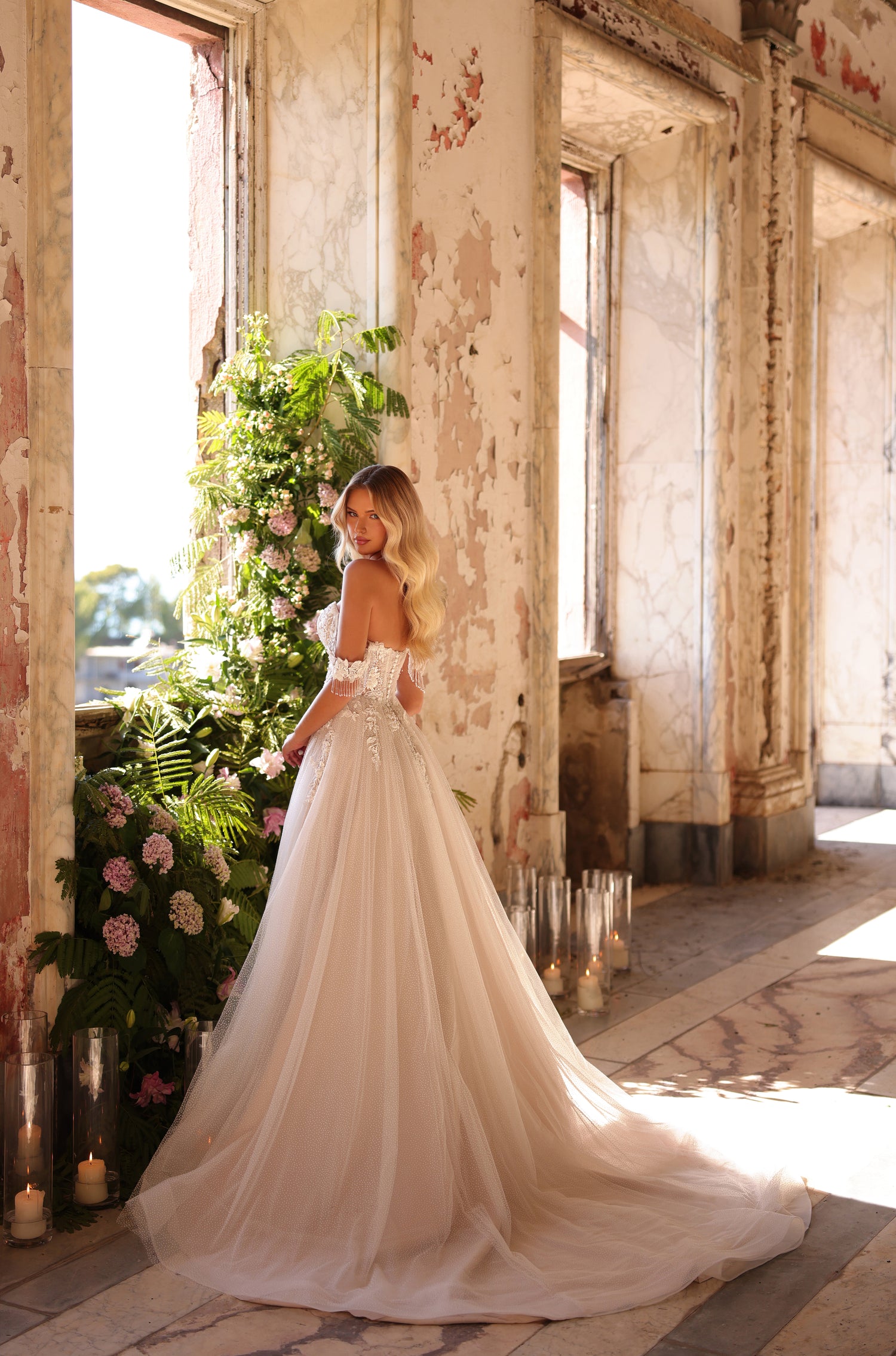 A bride in a lace off-shoulder wedding gown poses in a sunlit, distressed room with floral and candle decor.