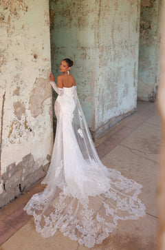 A bride in an off-shoulder lace wedding gown with a long train stands in a weathered, textured room.