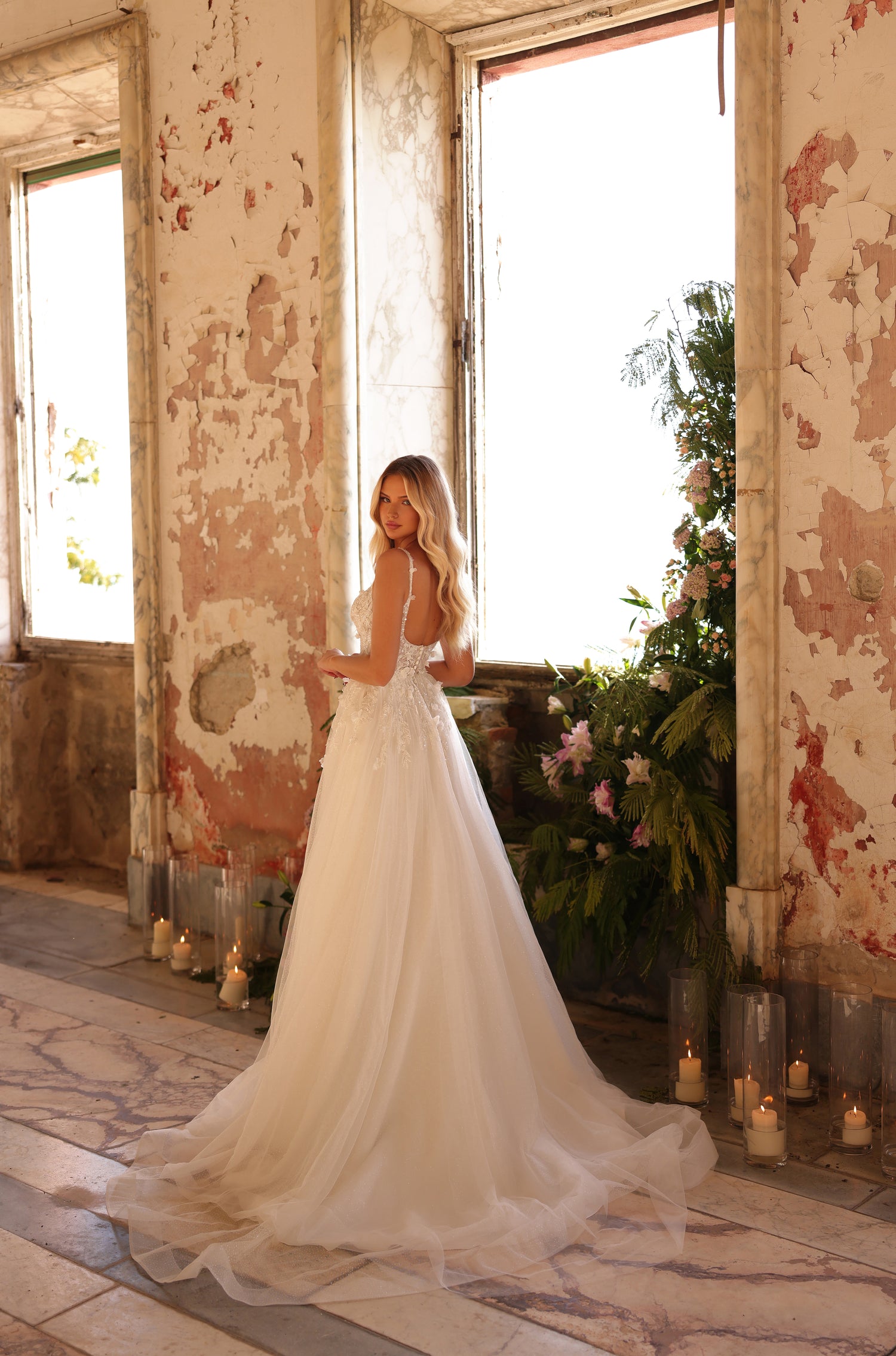 Bride in flowing white wedding gown stands by window with candles, near lush floral arrangement in distressed room.