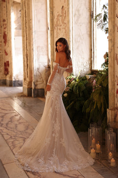 A bride in an off-shoulder lace mermaid wedding gown stands in a vintage, plant-filled room with candles.