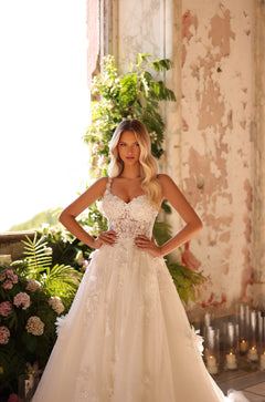 A blonde bride in an embellished lace ballgown stands confidently amid greenery and soft candlelight.