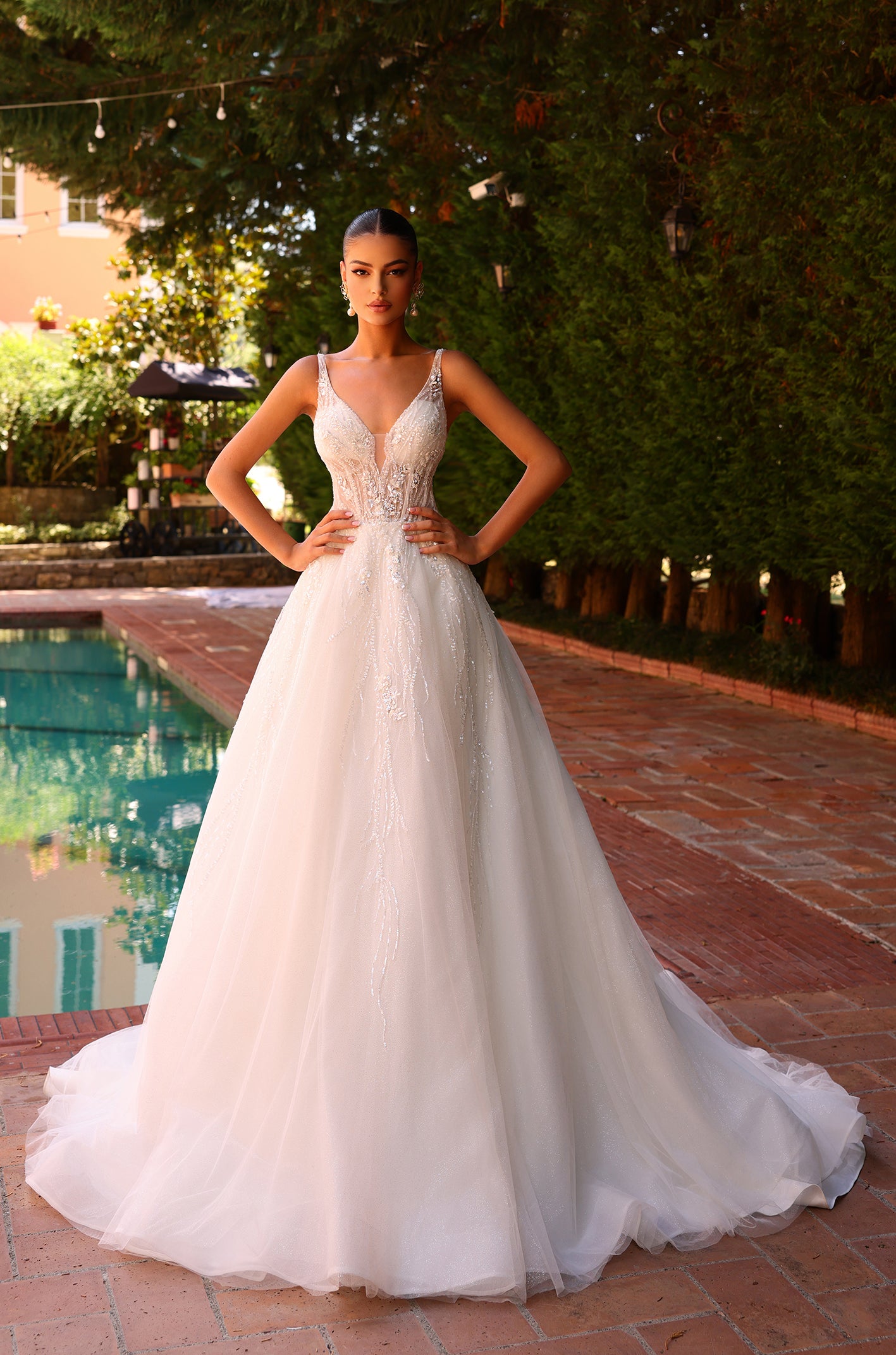 A model wears a white beaded A-line wedding gown by Style Daisy, standing poolside with hands on hips.