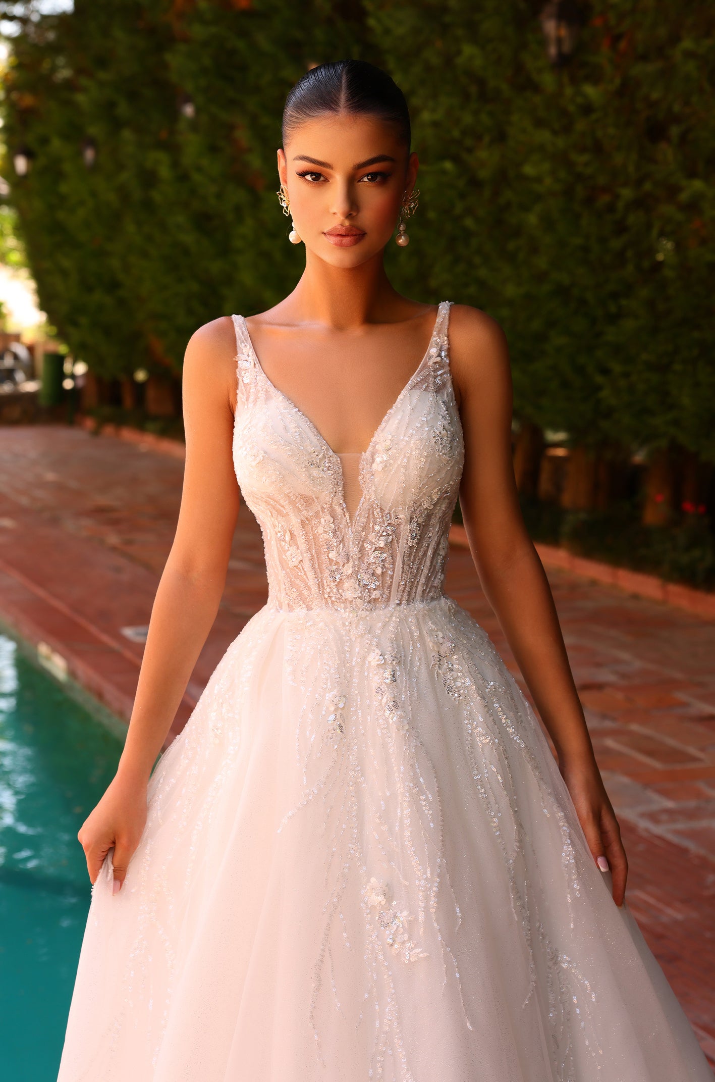 Model in shimmering white wedding gown with deep V-neckline stands poolside in elegant pose.