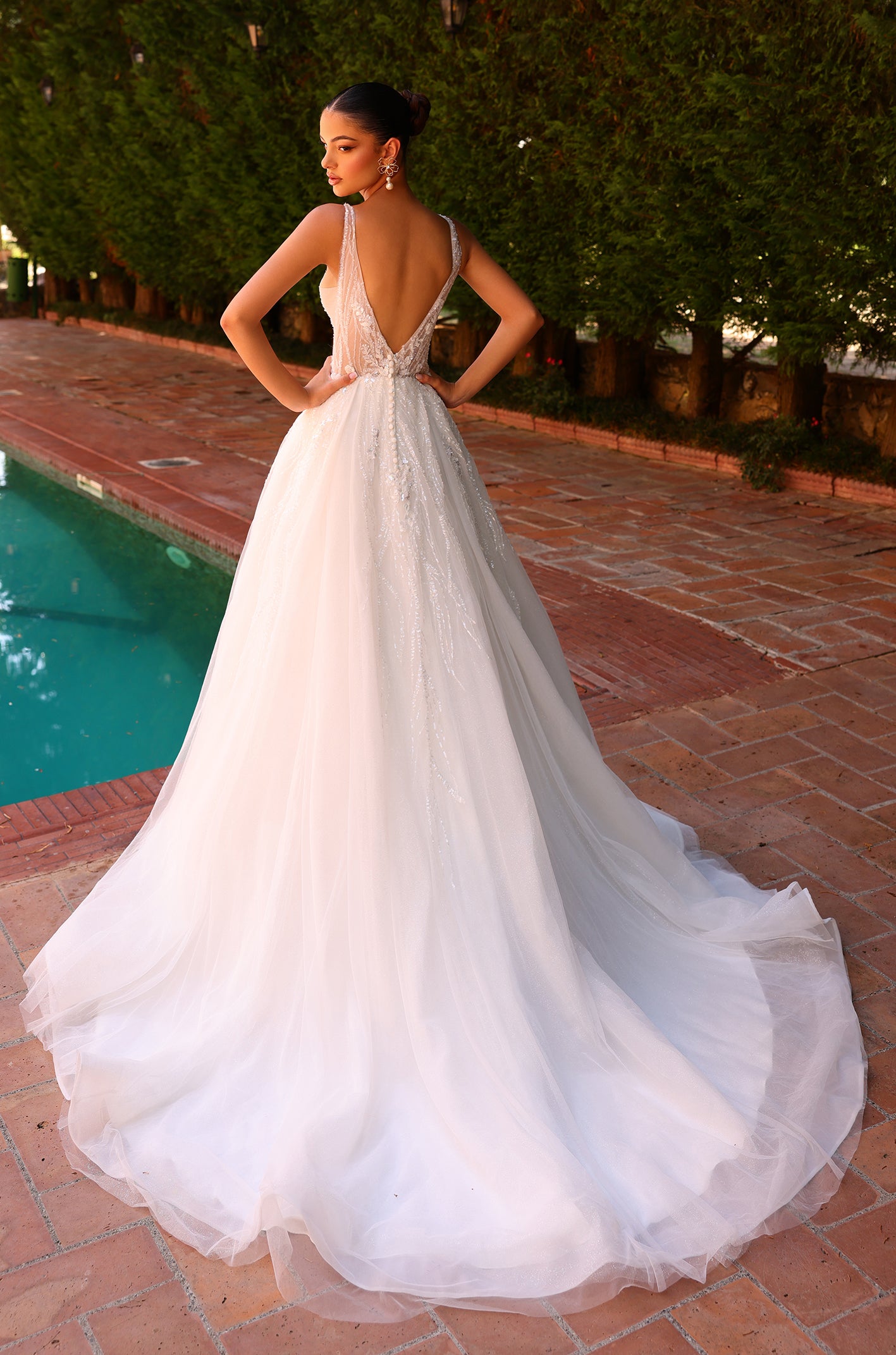 A white wedding gown with a deep V-back, sparkling embellishments, and a flowing tulle skirt, posed by a pool.