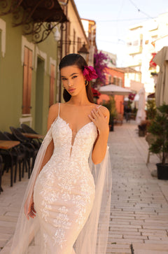 A bride in a white lace gown with a deep V-neckline stands on a charming street with a bright pink flower in her hair.